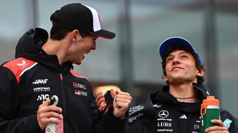 Getty Images Ollie Bearman's standing on the left with Kimi Antonelli on the right. They are both laughing and turned towards each other. They are holding a water bottle each. Both drivers are wearing black team jackets. Antonelli is wearing a blue Mercedes hat and Bearman is wearing a red, white and black Haas cap. 