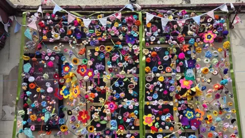 Wem Town Council A close up of the flowers on the pub. They are attached to netting and are made from an array of rainbow and colourful wool. There are sunflowers, daisies, pansies and more.