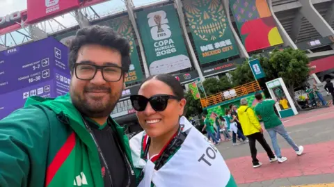 Aaron Vieyra A man and a woman taking a selfie in front of a football stadium. The man is wearing a green jacket and the woman has a white and green flag tied around her like a cape