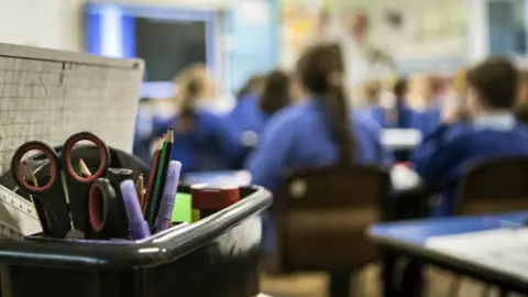 The picture shows a classroom scene with the pupils blurred out. The foreground is in sharp focus and shows a black plastic caddy filled with classroom supplies. Inside the caddy there are several pairs of scissors, a ruler, coloured pens, pencils, glue sticks and a sheet of graph paper leaning against the side. 
