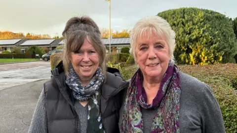 Ben Schofield/BBC Gloria Flack and Jean Bell standing next to each other and looking directly down the camera. They are both standing outside, in a residential street, with bushes and bungalows in the background. Gloria, on the left of the picture, has dark hair pulled into a high pony tail. She is smiling, with her teeth showing, and is wearing a dark gillet over a grey ribbed jumper and a black, blue and white silk scarf. Jean is on the right of frame. She has light blonde hair and is wearing a grey jumper and a patterned purple and green silk scarf. It looks like a bright day.  