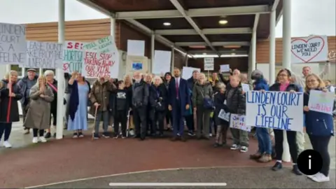 About 40 campaigners, including the Eastbourne MP in the centre, stand in a semi-circle holding various banners including "Linden Court is our lifeline" and "East Sussex County Council stop taking away our vital services".