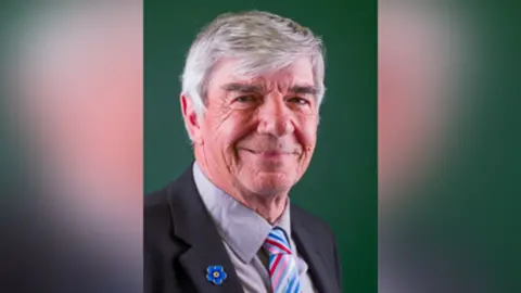 Peter York has short white hair. He is standing against a green background. He is wearing a dark coloured suit with a grey shirt and multi-coloured striped tie. He has a blue flower pin on his lapel.