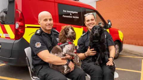 WMFS A man and a woman in black fire service uniforms sit together with a dog on each of their laps. The man, sat on the left, holds a brown and white dappled cocker spaniel, while the woman holds a black cocker spaniel. A fire vehicle is parked behind them.