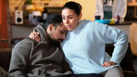 BBC/Jack Barnes/Kieron McCarron A still showing Ravi with his eyes closed, leaning on Priya, who is wearing a blue hoodie. They are both sitting on a sofa.
