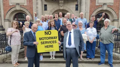 LDRS A large group of men and women stand outside a town hall holding a yellow sign that reads: No motorway services here