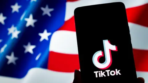 A TikTok logo is displayed on a smartphone with the U.S. flag in the background