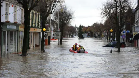 Owen Humphreys / PA Media Members of the RNLI are in a small boat, travelling down a high street that has been submerged in brown flood water. The water comes up to just under the front window of a blue van on the right. Black and white poles, marking a zebra crossing, rises out of the water, as do trees.