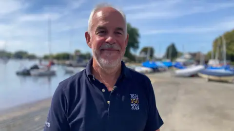 A man in a blue polo shirt stands in front of a harbour on a sunny day. He is smiling.