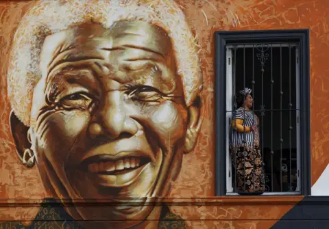 ESA ALEXANDER / REUTERS A large sepia-toned mural of a smiling Nelson Mandela covers the side of a building. A woman stands and smiles in one of the windows as she poses for a photo.