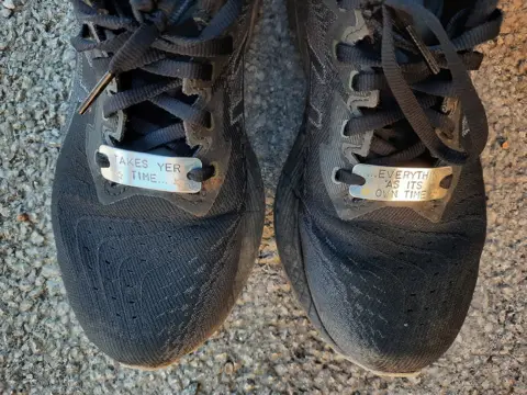 Elizabeth Rees-Gilbert The toes of two black running shoes. Each has a silver plaque which are laced to the shoes. One reads: Takes yer time. The other says: Everything 'as its own time. 