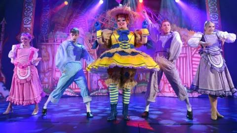 Five people on stage singing and dancing in pantomime costumes.