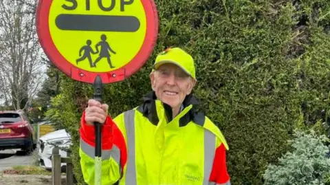Reg dressed in high-vis uniform, coat and gate - holding a lollipop sign reading 'stop' with the highway code symbol for children/ school
