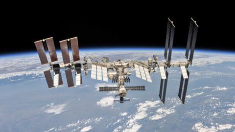 The International Space Station photographed by an astronaut with earth in the background. It has a long reflective surface with lots of solar panels sticking out from it.