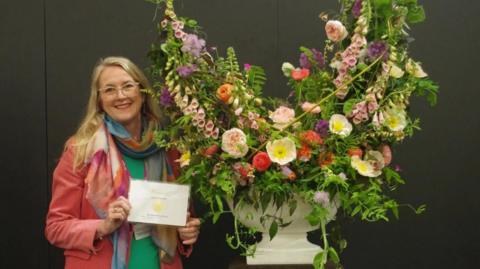 Nicola standing next to her flower arrangement holding a gold medal certificate