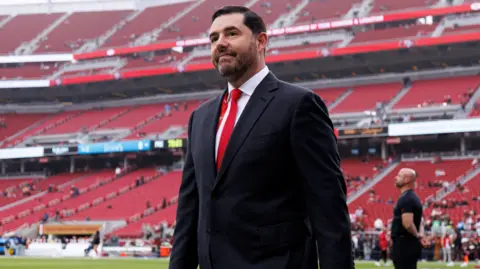 Getty Images Jed York stands on the field prior to an NFL football game against the Tennessee Titans at Levi's Stadium.He is wearing a dark suit, white shirt and red tie.