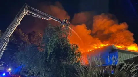 A grey building with flames and smoke billowing from the roof. There is a metal extendable ladder to the right, with a firefighter directing a jet of water at the fire. There are trees and grasses in the foreground.