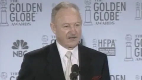 Gene Hackman pictured in 2003 wearing a dark suit and white striped tie in front of  Golden Globes backdrop