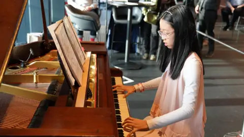 A young girl plays a piano. She has shoulder-length black hair with a thin hairband. She is wearing a pink outfit. She is playing a brown wooden piano and some people are in the background watching on.