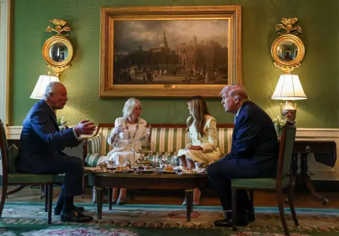 Pool/Getty Images King Charles, Queen Camilla, First Lady Melania Trump and US President Donald Trump sit around a low table on which cups, saucers and afternoon tea snacks are placed. King Charles is gesturing with his hands as he speaks to Trump, while Queen Camilla is doing the same as she converses with Melania. They are sat in a room decorated with green walls, gold-framed mirrors and a large framed painting.