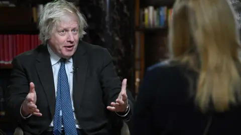 Former prime minister Boris Johnson speaking while holding out both hands. In the foreground, out of focus, we see the back of a woman with blonde hair.