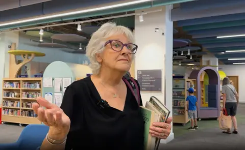 A woman with grey hair, glasses and a black top, carrying some books. 