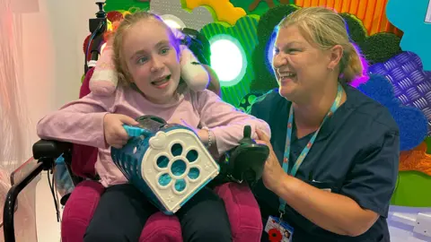 Edith Williams with Nurse Lucy Reilly in the sensory room. Edith is in a motorised wheelchair and holds a toy. Lucy is kneeling beside her, holding her arm. Both are smiling broadly and both have tied-back blonde hair.