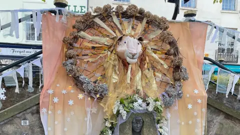 A decoration over a well includes a peach coloured fabric background, a model of a lion surrounded by sun rays, and snowflakes on the cloth. There is a wreath made of twigs and a floral garland.