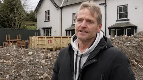 A man with short, light brown hair, swept to one side, and light stubble, stands by a pile of earth in front of a house with several windows amid white walls. He wears a grey hoodie under a black coat.