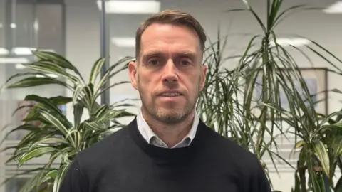 A man in a black jumper and striped white shirt stands in front of several plants. He has brown hair and stubble.