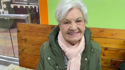 Spencer Stokes/BBC A woman with white hair and a green coat and pink scarf in a cafe at a shopping centre. 
