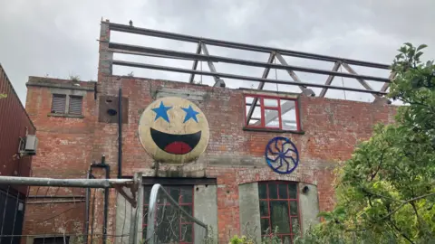 A fenced-off, derelict building. Its roof has been completely stripped and just the bare structure visible. There is a big smiley face on one wall, which is yellow with blue stars as eyes.