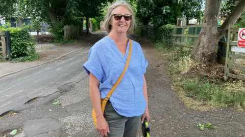 A woman wearing a light blue short-sleeved top and grey trousers stands at the edge of a potholed road near a tree. She has an orange messenger-style bag and is wearing sunglasses and holding a dog lead.