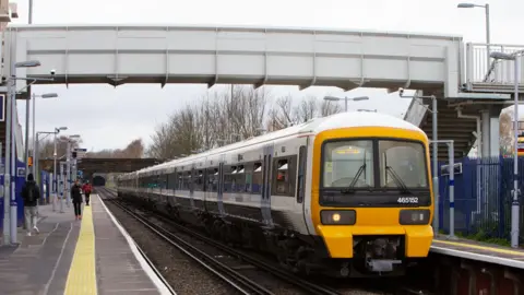 A white Southeastern train with a yellow front and blue doors at a station with a footbridge connecting two platforms