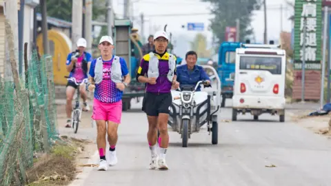 Jo Lodder Two men in sport clothing run on a busy road with vehicles driving past. They are both wearing caps, shorts and running vests. 