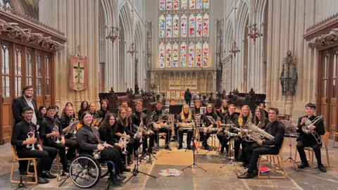 Ianto Williams Ianto Williams (far left) with the rest of the universities brass band. They are performing in a church hall and are all looking at their camera holding their instruments. 