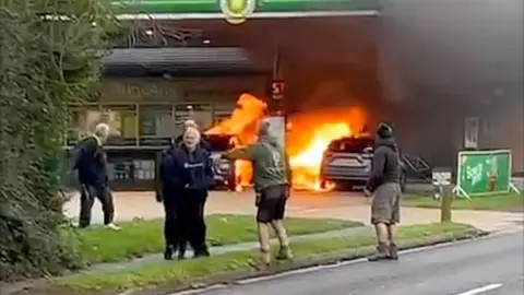 Five people standing on a path by a petrol station forecourt where behind them two vehicles are on fire and there is a large amount of black smoke.