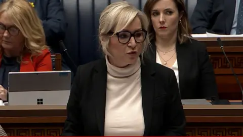 BBC Michelle O'Neill speaking in the Stormont assembly. She has shoulder-length blonde hair and is wearing a dark jacket with light coloured top.