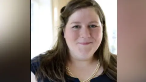 Supplied Laura Courty has straight brown hair, which is half up. She is wearing a navy blue short-sleeved top and two strings of pearls. Her eyes are blue. 