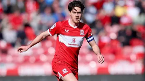 Hayden Hackney, with the ball at his feet and wearing the captain's armband, playing on a sunny afternoon for Middlesbrough