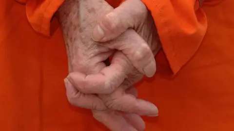 Generic image of clasped hands, they belong to an older person, who is wearing an orange long sleeve top. Their face is not seen.