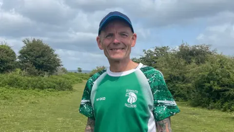 A man in a blue cap, smiling at the camera, wearing a green Plymouth Falconers top. There is green grass and bushes behind him. The sky is filled with white clouds with small bits of light blue.