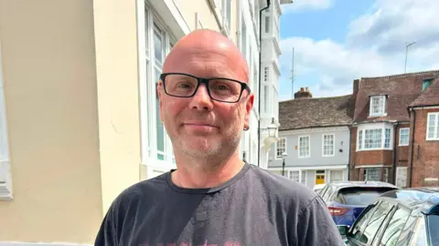 A man wearing glasses and a t-shirt. He is stood in a street.