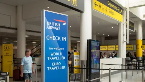 Getty Images A check in area at London Gatwick has a sign for British Airways. Staff are being a desk and a man in a T-shirt is near a sign that says self check-in. There is a screen near the check-in desks. A yellow sign overhead points to Departures.