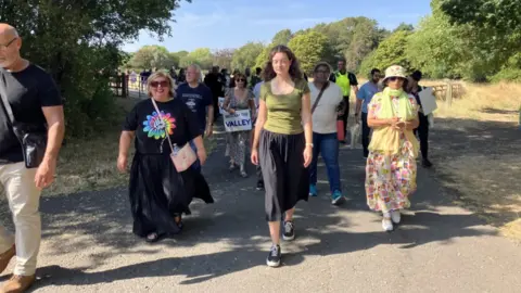 A group of around 20 people walk along a paved path through a park or nature area on a sunny day. Trees line both sides of the path, and several people are carrying signs, including one that reads “Reclaim the Valley.” The group is spread across the width of the path, with open countryside and greenery visible in the background beneath a clear blue sky.