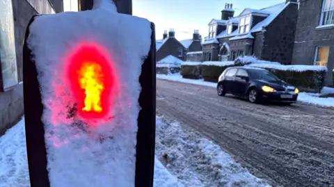 Una señal de tráfico con forma de "hombre rojo" cubierta de nieve, con un coche con las luces encendidas, sobre una carretera cubierta de nieve.