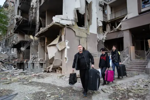 Three people carrying suitcases leaving a partially destroyed building