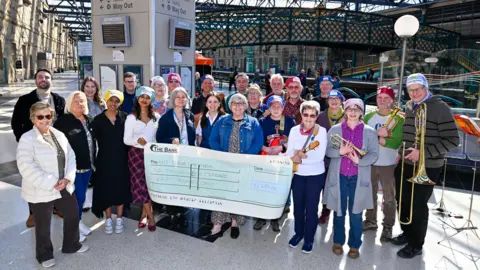 Supplied A group of people stand together inside a railway station beneath a glass roof, holding a large presentation cheque. Station signs, information boards, and rail platforms are visible in the background, and several people are holding brass instruments.