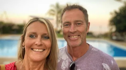 A man and a woman smile. Behind them is a blurry swimming pool. 