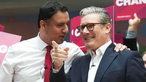 Anas Sarwar standing next to Sir Keir Starmer at a Labour Party event. Sarwar is wearing a white shirt and red tie and has his hand on the shoulder of Starmer, who is wearing a white shirt, a blue jacket and black-rimmed glasses.